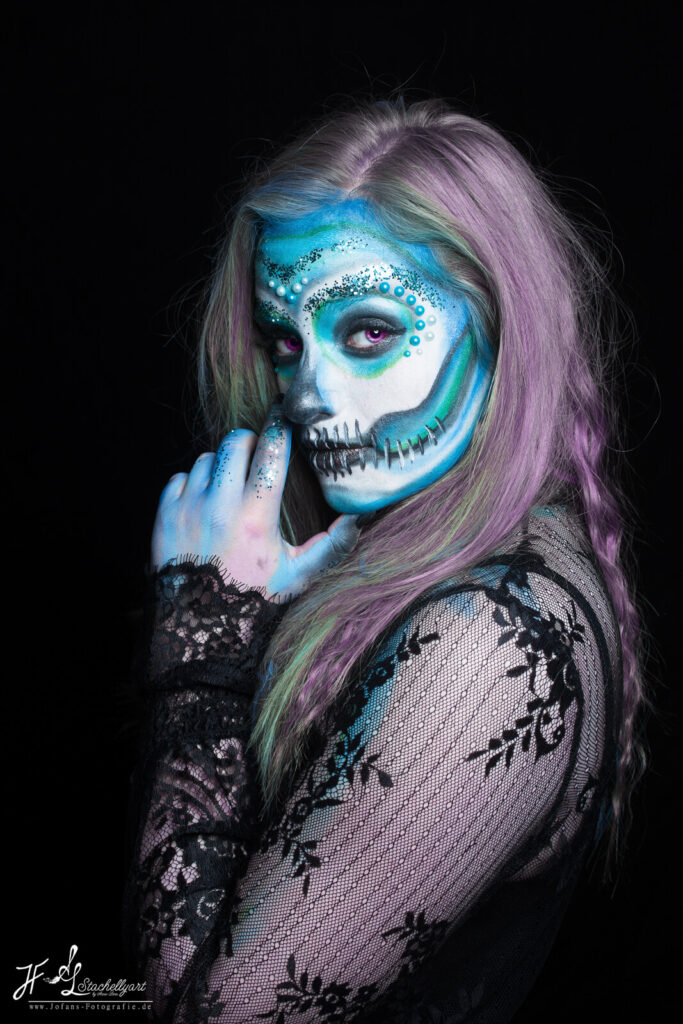 Mädchen mit Facepainting eines blauen Sugarskulls mit Perlen schaut über die Schulter in die Kamera. 